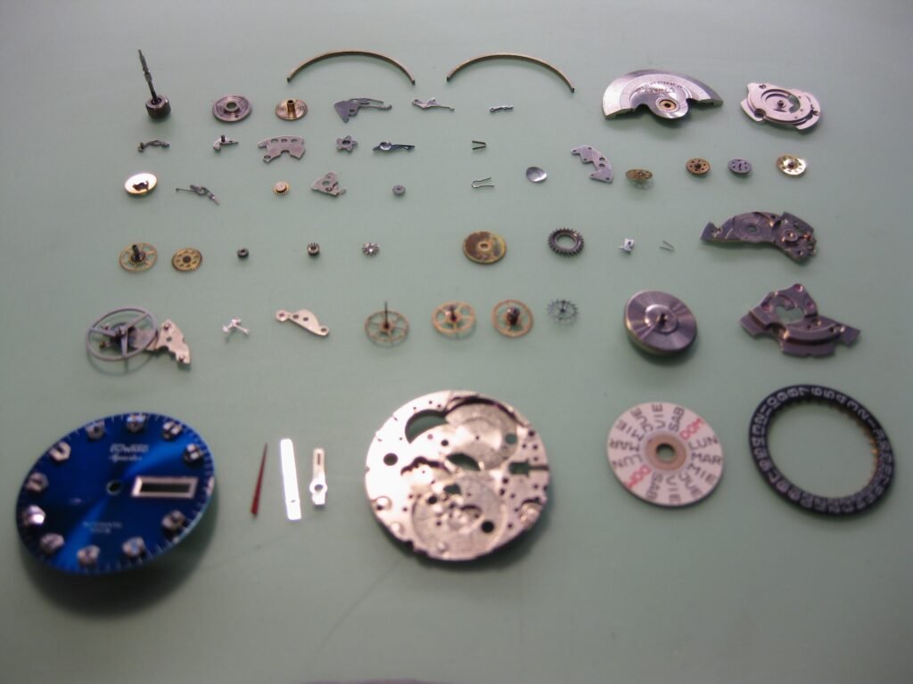 Movimiento de reloj mecánico completamente desmontado, con piezas metálicas dispuestas en filas ordenadas sobre una superficie verde clara.