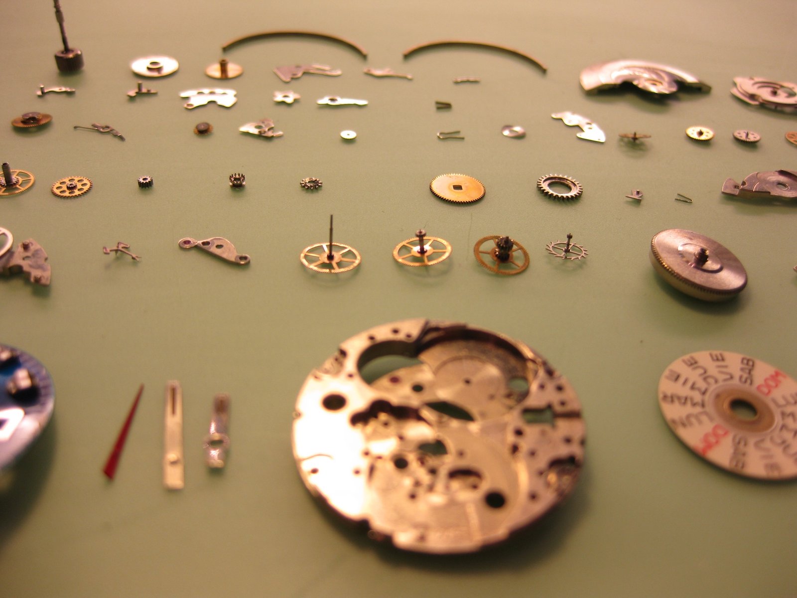 Componentes de un reloj mecánico desmontado, organizados en filas sobre una superficie verde clara. Se distinguen engranajes, ruedas dentadas, ejes, puentes y la platina principal en primer plano, con enfoque selectivo que resalta las piezas centrales.