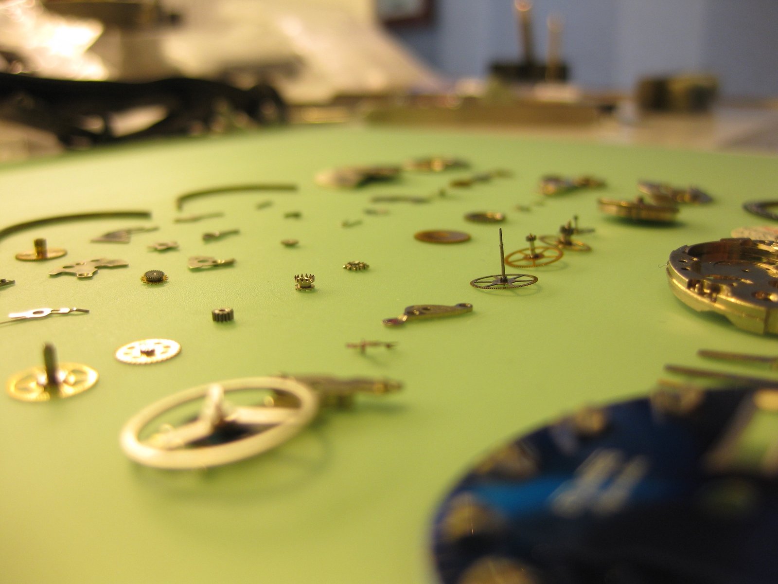 Piezas de un reloj mecánico desmontado distribuidas sobre una superficie verde clara, vistas en perspectiva baja. Se aprecian engranajes, ejes, ruedas y pequeños componentes metálicos, con fondo desenfocado que sugiere un entorno de taller.