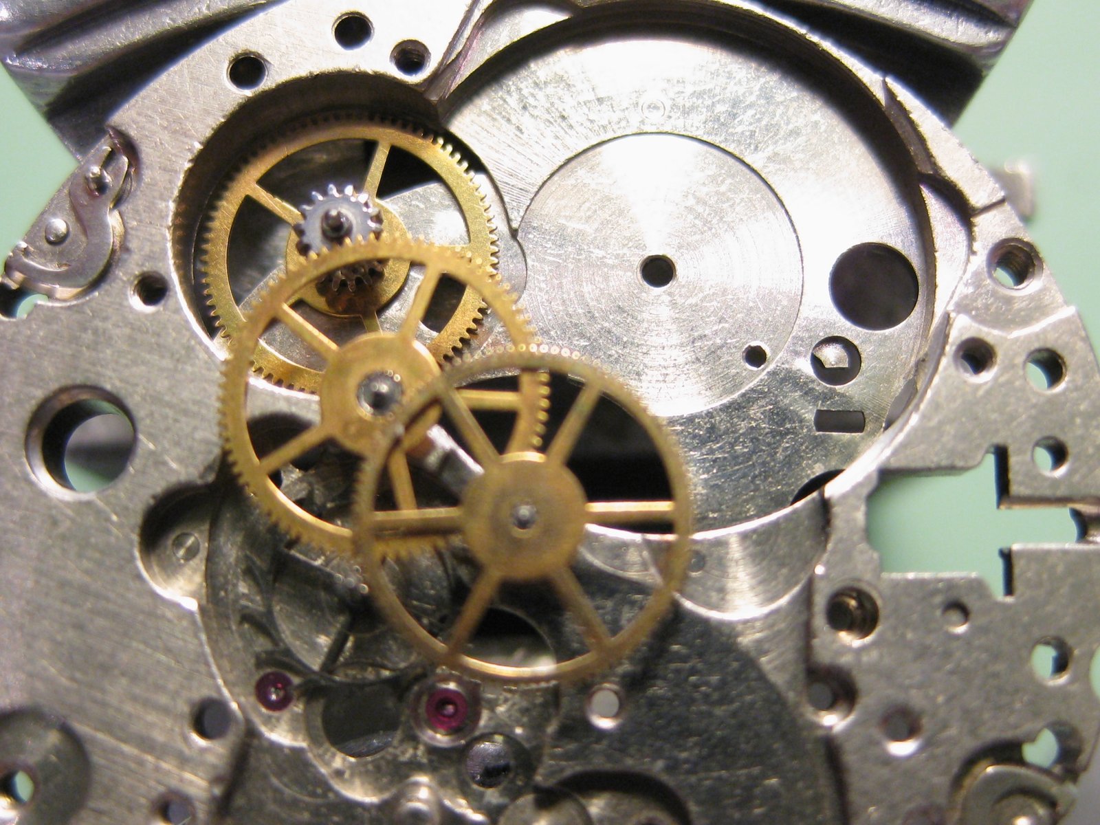 Detalle macro de engranajes dorados en un movimiento de reloj automático ETA 2789: ruedas del tren de marcha con dientes finos, pivotes con joyas rubí rojas visibles y tornillos de regulación, sobre placa metálica plateada con perforaciones.